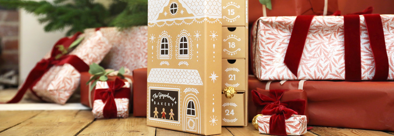 Gingerbread House Style Advent Calendar With Filled Drawers Visible Sat in Front of a Christmas Tree Surrounded by Presents