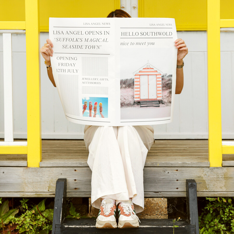 Founder, Lisa Angel Holding A Newspaper In Front of Beach Hut on Southwold Beach