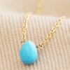 Close Up of Semi-Precious Turquoise Stone Teardrop Pendant Necklace in Gold on Beige Fabric