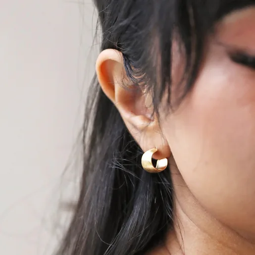 Close Up of Model Wearing Brushed Wide Gold Huggie Hoop Earrings in Gold with Cream Background