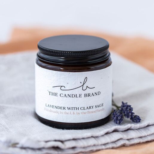 The Candle Brand Lavender and Clary Sage Scented Candle on cloth with lavender flower next to it