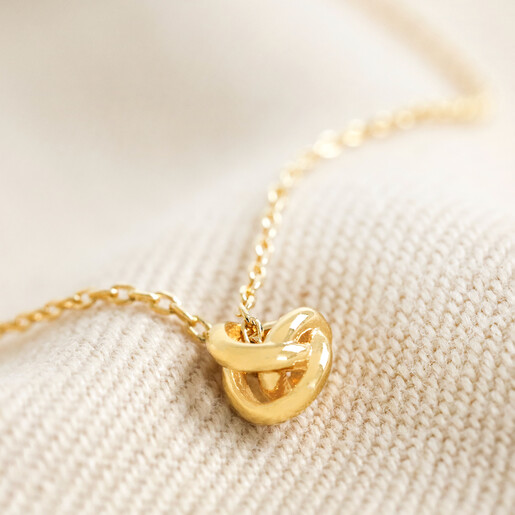 Close Up of Estella Bartlett Knot Pendant Necklace In Gold on Beige Fabric
