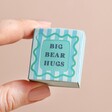 Model holding the Tiny Matchbox Ceramic Bear Token packaging between fingers against neutral background