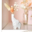 Tiny Elephant Bud Vase on white shelf with flowers inside and vase in background