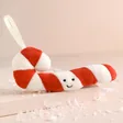 Jellycat Plush Festive Folly Candy Cane Decoration lying amongst fake snow on pink surface