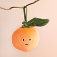 Jellycat Plush Festive Folly Clementine Decoration hanging from tree branch against neutral background