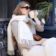 Model in sunglasses drinking coffee while wearing the cream Personalised Embroidered Cashmere Blend Scarf