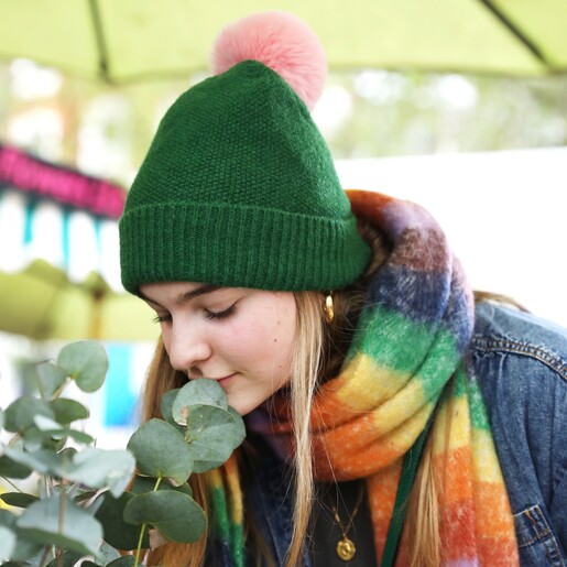 Model smelling shrubbery wearing Vibrant Rainbow Striped Winter Scarf and green bobble hat