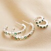 Set of 2 Green Crystal Huggie and Hoop Earrings in Silver laid out on top of neutral coloured fabric