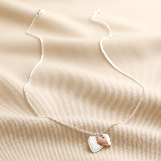 Mixed Metal Textured Double Heart Necklace in Silver laid out full length on top of beige coloured material