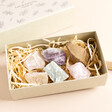 A selection of colourful stones inside the East of India Crystal Matchbox Set. Placed on a beige background with wooden shavings inside the box.