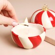 Red and White Stripy Ceramic Jar Candle in the Shape of a Bauble Being Lit With a Match