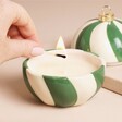 A Green and White Striped Ceramic Jar Candle With The Wick Being Lit By a Match