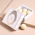 Opened Box of White Chocolate Champagne Filled Truffles With White and Gold Lid Raised on a Neutral Background