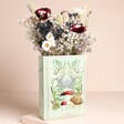 A Pale Green Ceramic Vase in the Shape of a Book With a Woodland Illustration on the Front Filled With Dried Flowers