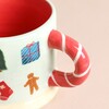 Close up of mug, featuring red candycane handle and festive print such as gingerbread and a present. Beige background.