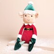 Jellycat Eldo Elf Soft Toy on a beige background with a green hat and red outfit