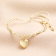 A charming gold beaded bracelet on a beige cord with a dangling heart charm in gold.