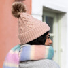 A Chunky Pastel Pink Knitted Hat With a Brown Fluffy Bobble Being Worn by Model With Pink and Blue Stripe Scarf