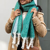 A Model Wearing a Green and Blue Tonal Winter Scarf with Chunky White Braided Tassels Styled With a Tan Trench Coat