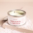 A Metal Tin With a Cream Label Filled With a Cinnamon & Orange Scented Wax Candle on a Neutral Background