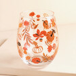 A stemless glass with pumpkins, mushrooms, leaves and acorns illustrated across it in shades of red, browns, and oranges.