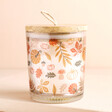 A vanilla scented candle with a wooden lid and an autumnal external design featuring pumpkins, flowers, mushrooms and acorns