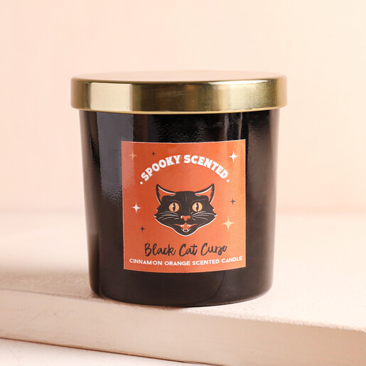 A black glass candle with a gold lid and an illustration of a black cat on an orange background