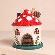 A charming mushroom house with a removable green base. The house has a snail with a happy face on the roof, two windows and a wooden door