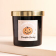 A candle made with black glass and a gold lid with a pumpkin carving illustration