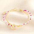 A dual layered bracelet with a gold beaded layer and a pink and white agate beaded layer with a doodle style heart charm