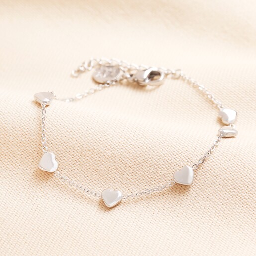Stainless Steel Heart Charm Bracelet in Silver on a Beige Background. A dainty heart charm bracelet with silver stainless steel hearts seperated by a trace chain and lobster fastening.