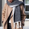 A Reversible Charcoal Coloured Blanket Scarf Worn Over one Shoulder on a Model in a Tan Coat