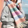 A Model Wearing a Pastel Coloured Blanket Scarf in Shades of Pink Green Blue and Orange With a Grey Coat