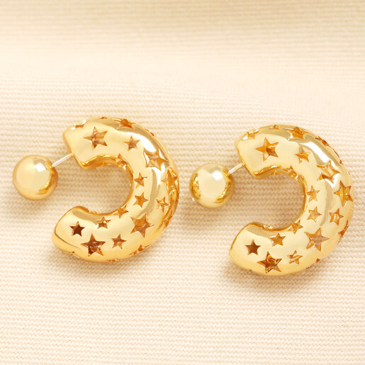 Gold chunky C-shape hoop earrings with cutout star details resting on a beige background