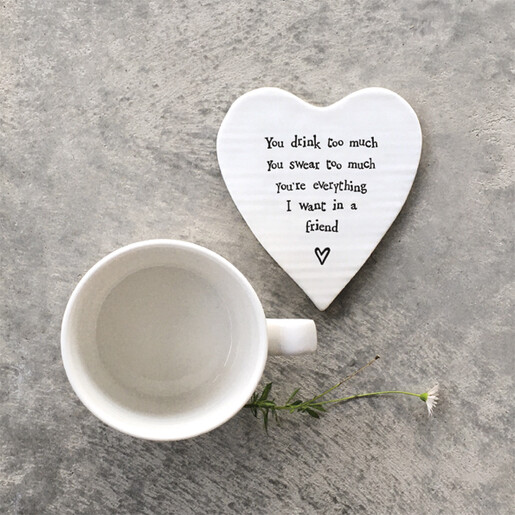 White ceramic heart coaster with humorous quote beside a white mug on a grey surface