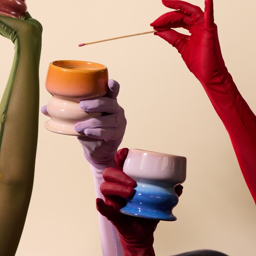 Gloved hands holding two colourful ceramic candle pots with a match held above