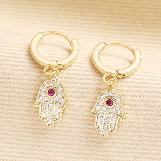 Pair of gold hamsa hand charm huggie hoop earrings with red crystal centers.