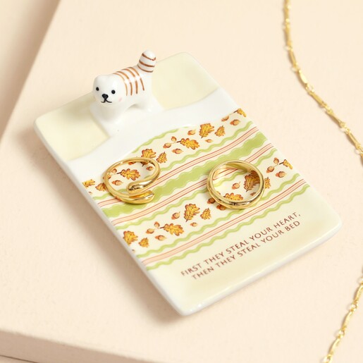 Ceramic bed-shaped cat trinket dish holding two gold rings on a bedside table