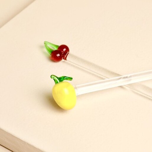 Glass lemon drink stirrer resting beside a glass cherry stirrer on a neutral surface