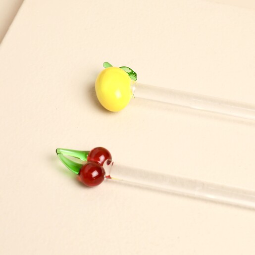 Clear glass drink stirrer topped with red glass cherries beside lemon stirrer