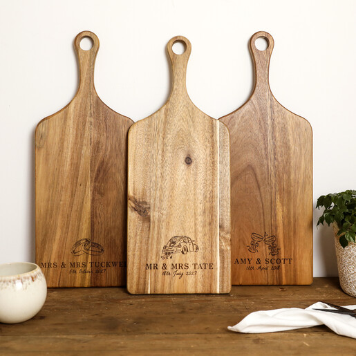 Three personalised acacia wood paddle serving boards showing different engraved wedding illustrations