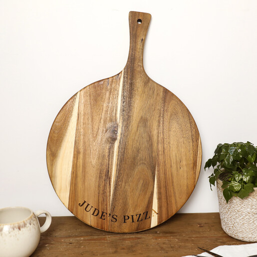 Personalised acacia wood pizza board paddle leaning upright beside a potted houseplant