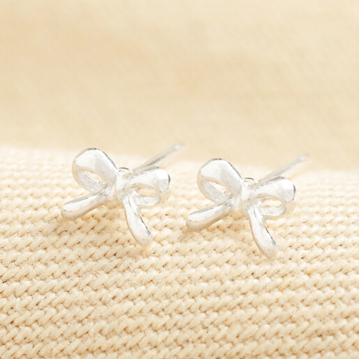 Dainty sterling silver ribbon bow stud earrings resting on a cream textured surface