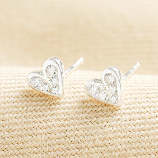 Pair of sterling silver crystal heart stud earrings on woven cream fabric.