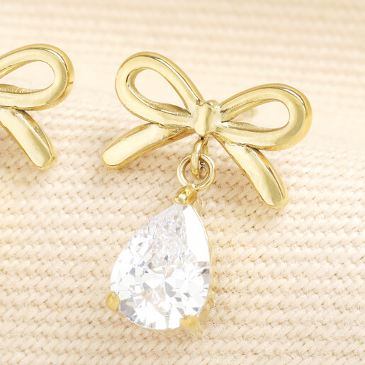 Close up of gold bow earring featuring a suspended teardrop faceted crystal charm