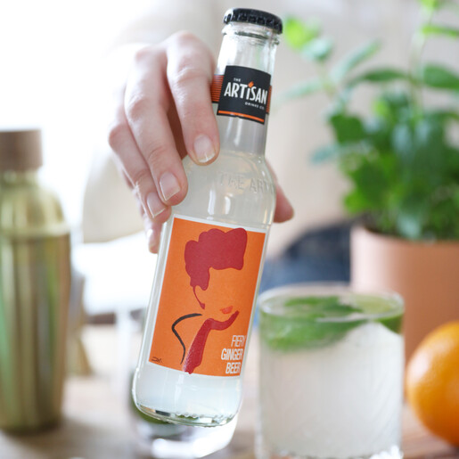 The Artisan Drinks Co. Fiery Ginger Beer Lisa Angel model holds the artisan drinks co fiery ginger beer with glass, plant and cocktail shaker in background