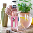model holds a bottle of pink citrus tonic with glass, plant and cocktail shaker in background