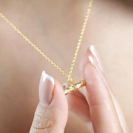Model Wearing Estella Bartlett Flower T-Bar Necklace in Gold and Holding Pendant Between Fingers