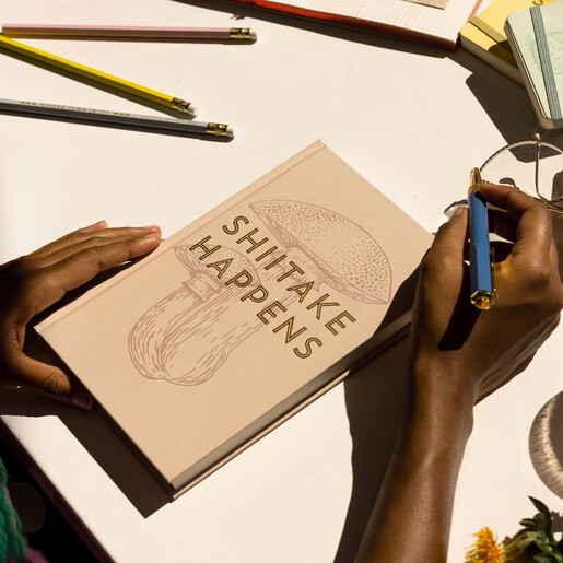 Model holding Designworks Ink Shiitake Happens Lined Notebook on desk with pen